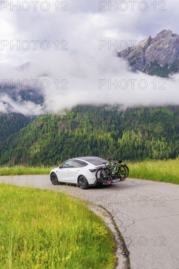 A car stands on a winding road with a view of misty mountains and green meadows, electric car on holiday, Dolomites, South Tyrol, Dolomites, Italy