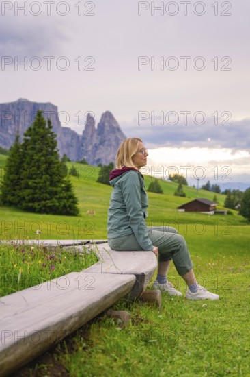 Woman sitting on a bench in a green landscape with mountains in the background, Alpe di Siusi, South Tyrol, Dolomites, Italy