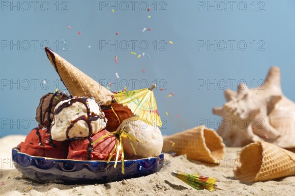 Various flavours of ice cream with chocolate sauce, crumble and parasol on sand with shells