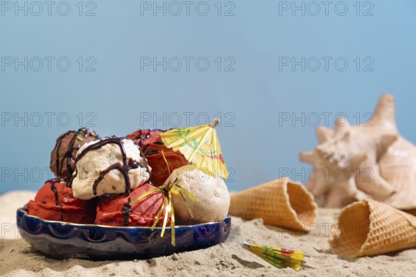 Various flavours of ice cream with chocolate sauce and parasol on sand with shells