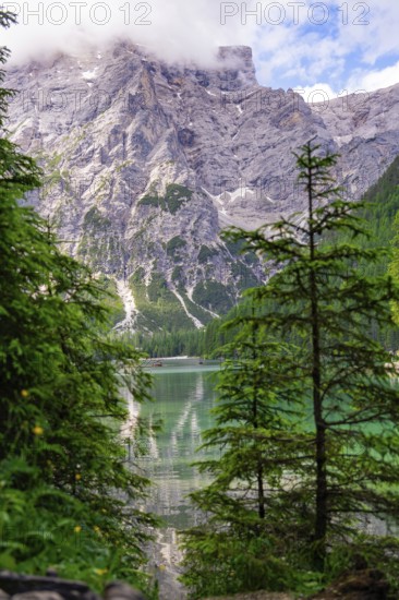 An imposing mountain reflected in a clear mountain lake, framed by green trees and a slightly cloudy sky, Lake Braies, South Tyrol, Dolomites, Italy