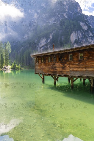 A wooden house stands on stilts above a clear, green mountain lake, surrounded by tall trees and majestic mountains, Pragser Wildsee, South Tyrol, Dolomites, Italy