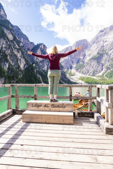 A woman stands with outstretched arms on a balcony and enjoys the view of mountains and a green lake, Lake Braies, South Tyrol, Dolomites, Italy