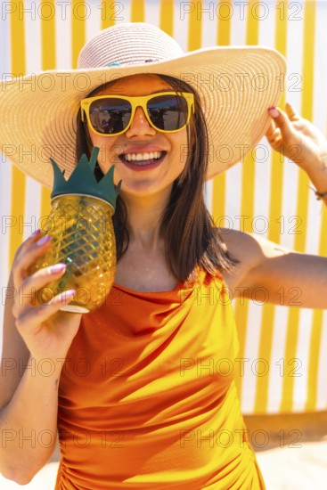 Happy woman wearing a straw hat and sunglasses holding a pineapple shaped cocktail in front of yellow and white striped background