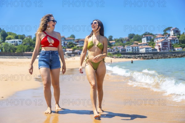 Two cheerful young women wearing bikinis and sunglasses are enjoying a walk on a sunny beach, creating a perfect summer vacation scene