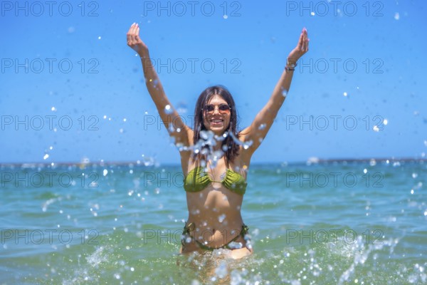 Young woman wearing sunglasses and a green bikini is raising her arms and splashing water in the sea, enjoying a sunny summer day at the beach