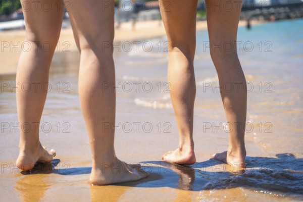 Two women standing barefoot on the wet sand of a sunny beach, enjoying the warm water and the summer holidays