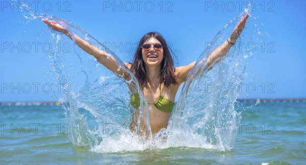 Young woman wearing stylish sunglasses and a vibrant bikini joyfully splashing in the clear ocean on a sunny summer day