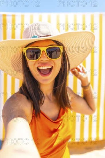 Young woman wearing sunglasses and a straw hat taking a selfie while smiling at the camera on a tropical beach