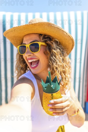 Happy tourist woman wearing sunglasses and straw hat taking a selfie at beach while holding a pineapple shaped cocktail