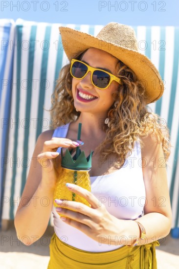 Young curly blonde woman wearing sunglasses and straw hat drinking juice at beach bar, enjoying summer vacation