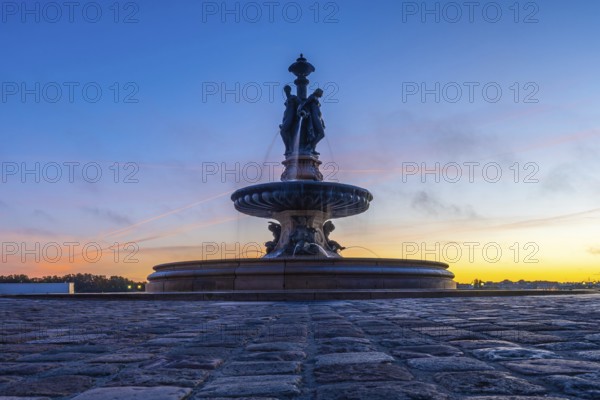 The three graces fountain in bordeaux, france, stands majestically against a vibrant sunrise sky, its waters flowing gracefully as dawn breaks over the city