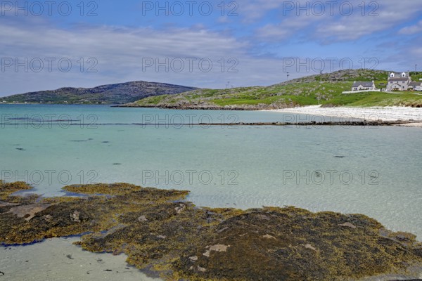 Clear water and a calm beach with hills and houses in the background under a partly cloudy sky, Eriksay, Hebrides, Scotland, United Kingdom