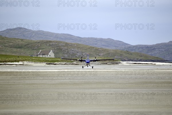 Aeroplane landing on a sandy runway on a beach with mountains and a building in the background, tidal, Loganair, Isle of Barra, Hebrides, Scotland, United Kingdom