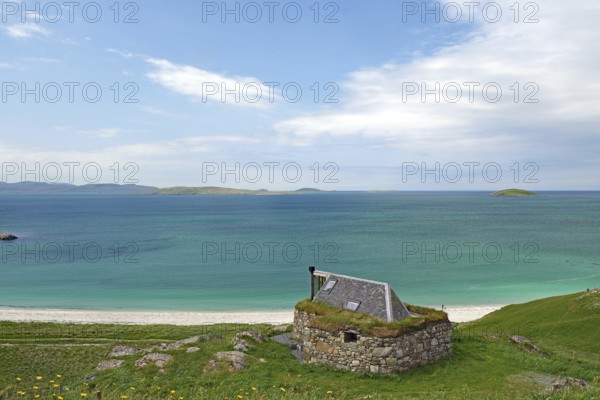 Stone cottage with grassy roof overlooking the vast sea and green hills under a blue sky, Eriksay, Hebrides, Scotland, United Kingdom