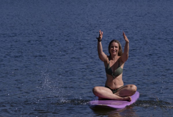Woman, blonde, in bikini, sitting on board, surfboard, surfboard, purple, back, from behind, splashing with water. Bathing lake, lake, Schwaltenweiher near Seeg, Ostallgäu, Allgäu, Swabia, Bavaria, Germany