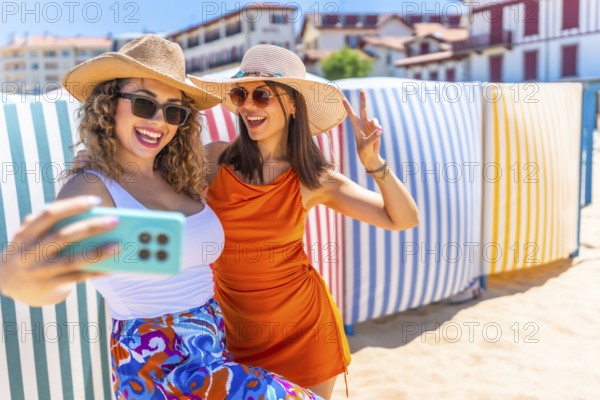 Two cheerful women wearing summer hats and sunglasses are taking a selfie with a smartphone on a sunny beach, gesturing peace signs in front of vibrant beach huts