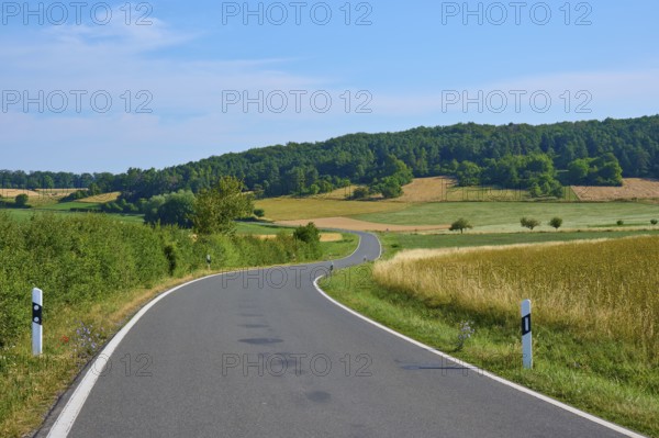 Curved country road through wide fields, surrounded by green forest and blue sky, summer, Neubrunn, Würzburg district, Bavaria, Germany