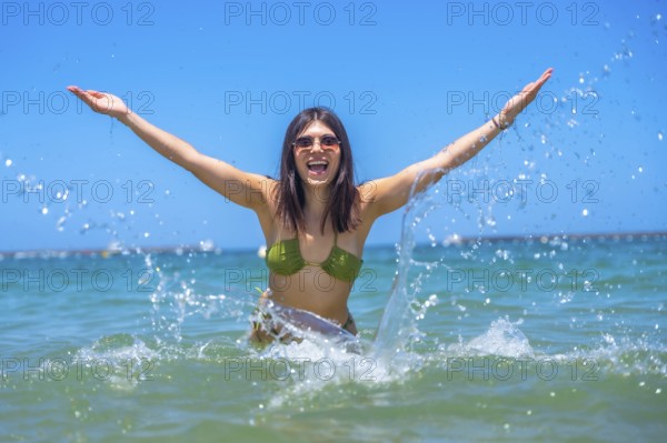 Young woman wearing sunglasses and a bikini is raising her arms and splashing water in the sea, enjoying her vacation