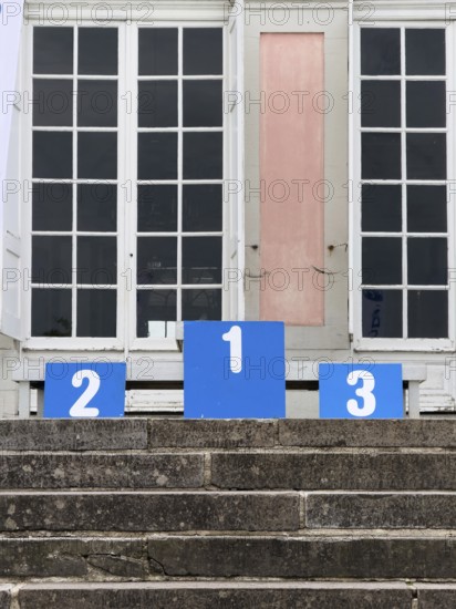 A winner's podium with the numbered places 1, 2 and 3, placed on a stone staircase in front of a building with large white double windows in Düsseldorf, Germany