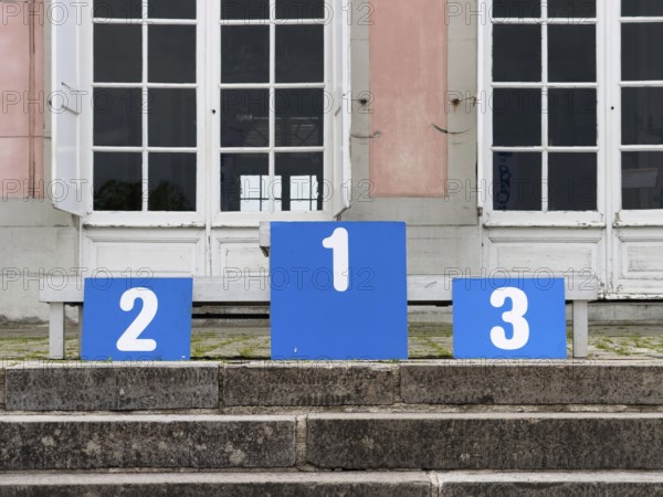 A winner's podium with the numbered places 1, 2 and 3, placed on a stone staircase in front of a building with large white double windows in Düsseldorf, Germany