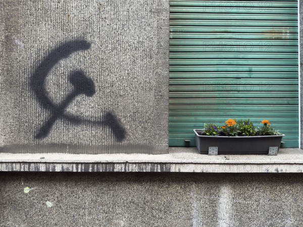 A hammer and sickle graffiti on a house wall next to a planted flower box in front of a roller shutter in Wuppertal, Germany