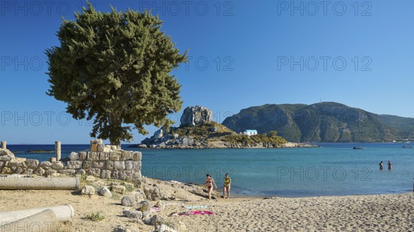 Family on the beach overlooking a green island and clear blue waters, Agios Stefanos Beach, Agios Stefanos Basilica, Agios Nikolaos Chapel, Kastri Island, Kos, Dodecanese, Greek Islands, Greece