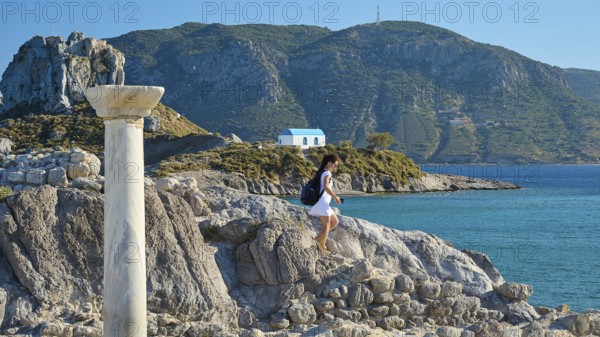 Person climbing on rock, ancient column in the foreground, island in the background, Agios Stefanos Beach, Agios Stefanos Basilica, Chapel of Agios Nikolaos, Kastri Island, Kos, Dodecanese, Greek Islands, Greece