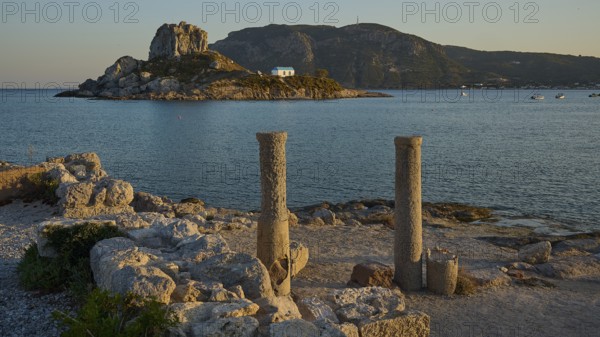 Two old columns in the foreground with a sunny island and blue water in the background, Agios Stefanos Beach, Agios Stefanos Basilica, Chapel of Agios Nikolaos, Kastri Island, Kos, Dodecanese, Greek Islands, Greece