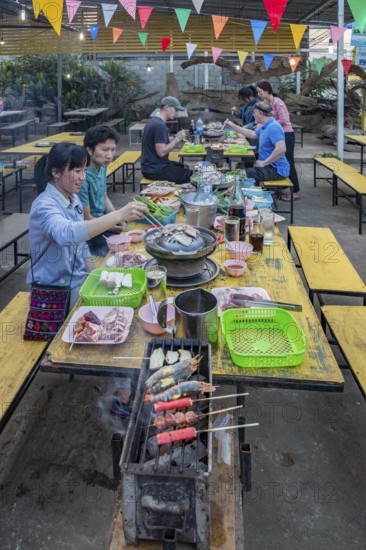 Thai man and woman cooking on a charcoal hot pot and grill at their table in a buffet style outdoor restaurant in Chiang Rai province of Northern Thailand
