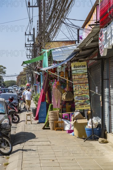 Tourists walk down narrow sidewalk filled with items for sale along Ban Du street in Chiang Rai, Thailand