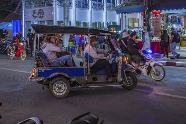 Motor scooter taxi with tourists merges into traffic on a busy downtown street outside the Night Market in Chiang Rai, Thailand