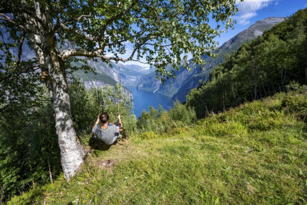 Young man swinging on a tree, view of idyllic fjord landscape of the Geirangerfjord at Blomberg Gård, near Geiranger, Møre og Romsdal, Norway