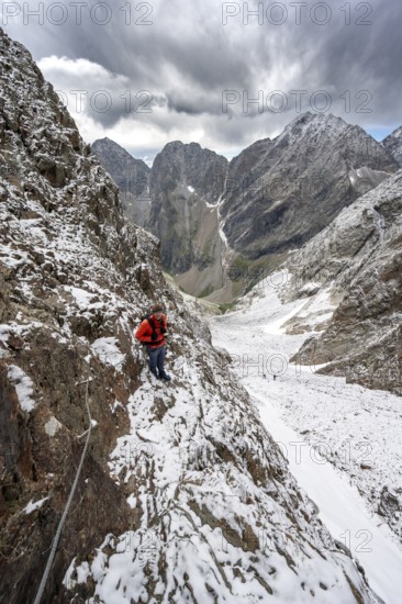 Mountaineer on a rope-secured path in a steep rocky mountain landscape with fresh snow in summer, behind mountain peaks Großer Friedrichskopf and Georgskopf, ascent to Hornscharte, Schobergruppe, Hohe Tauern National Park, Carinthia, Austria