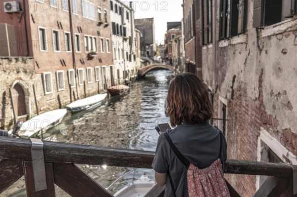 Young woman tourist with backpack using smartphone and enjoying view of picturesque canal, bridge, and historic buildings in venice, italy