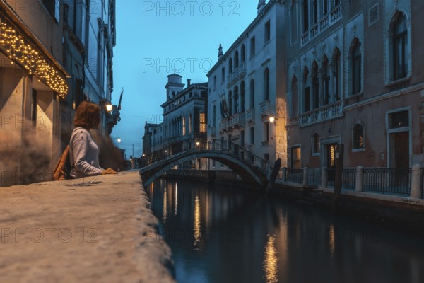 Young woman contemplating venice's canal at blue hour, with a small bridge and typical venetian buildings in the background