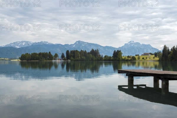 A tranquil lake with a wooden jetty, surrounded by mountains and trees under a cloudy sky