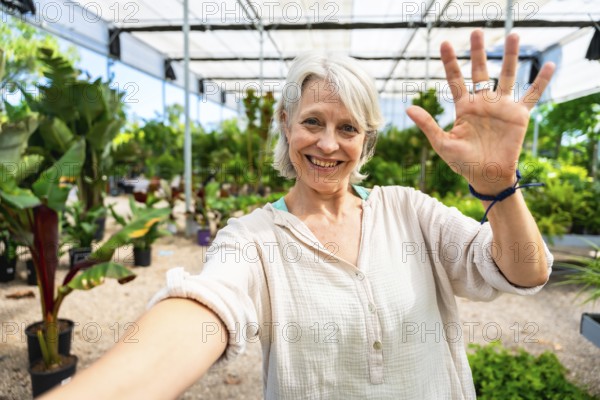 Happy gardener waving at the camera while taking a selfie, surrounded by plants in a greenhouse, enjoying her work