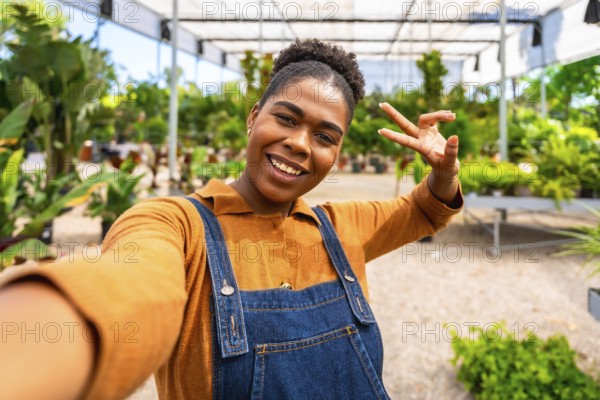 Happy young woman gardener taking a selfie and making a victory sign in a greenhouse full of plants