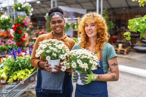 Two female garden center workers holding pots of white daisies smiling in a greenhouse or plant nursery