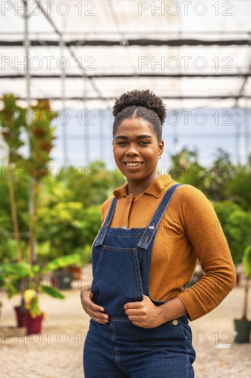 Confident agricultural worker smiling while holding her dungarees, surrounded by vibrant plants inside a well lit greenhouse