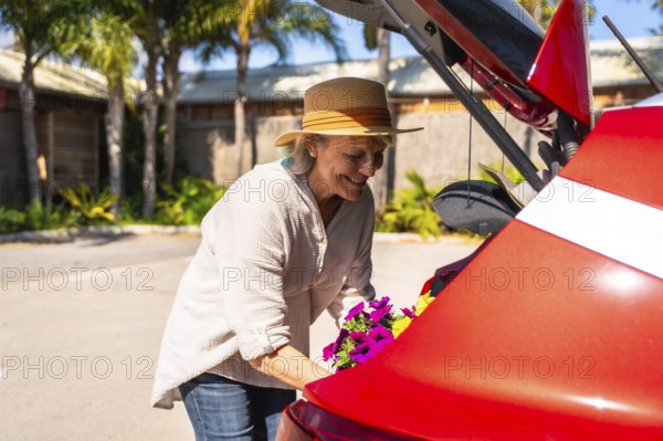 Happy senior woman carefully placing colorful petunias into the trunk of her red car, ready to bring them home