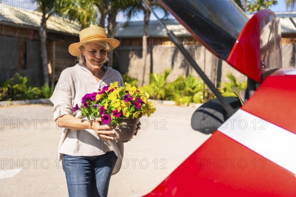 Happy senior woman carrying colorful petunias in a pot to the open trunk of her red car, ready for a sunny day trip