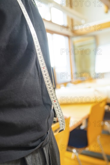 A man in a black T-shirt carries a tape measure over his shoulder in a light-coloured room with tables