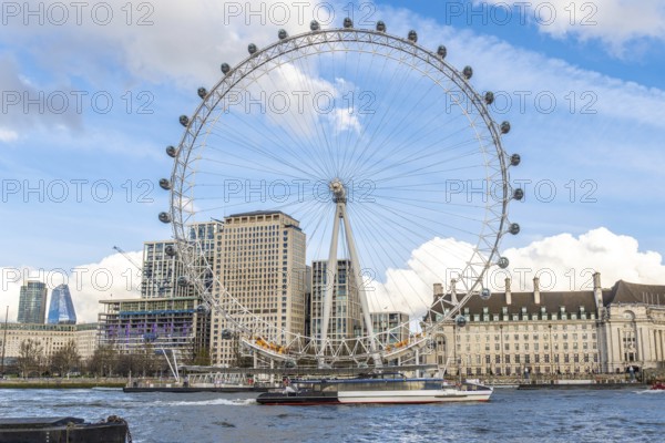 The london eye majestically stands tall on the banks of the river thames, with a boat sailing by and modern buildings in the background under a cloudy blue sky