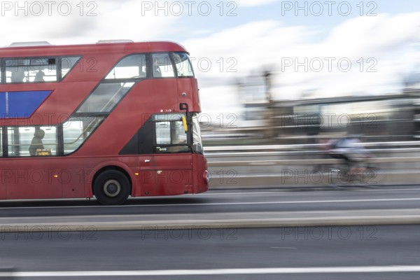 Red double decker bus driving on a london road with a cyclist passing by in the background, capturing the city's dynamic urban life