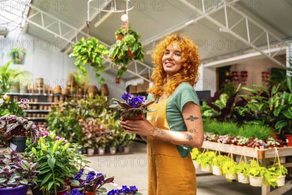 Young saleswoman holding a vibrant potted plant and smiling brightly in a lush greenhouse filled with an array of flourishing plants