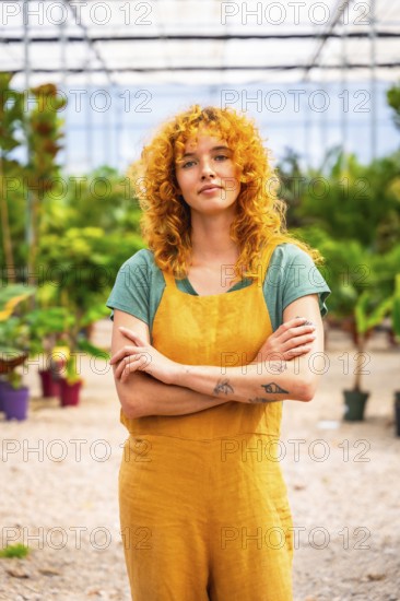 Young woman with curly red hair and tattoos wearing yellow overalls, standing with crossed arms in plant nursery greenhouse, looking confident