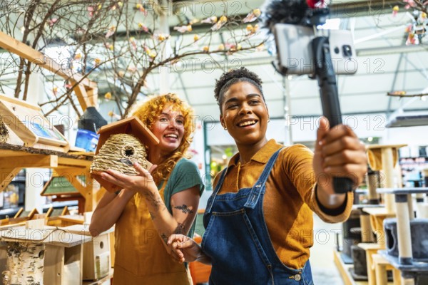 Two cheerful female shop assistants filming a vibrant video for social media while showcasing a charming birdhouse in their store