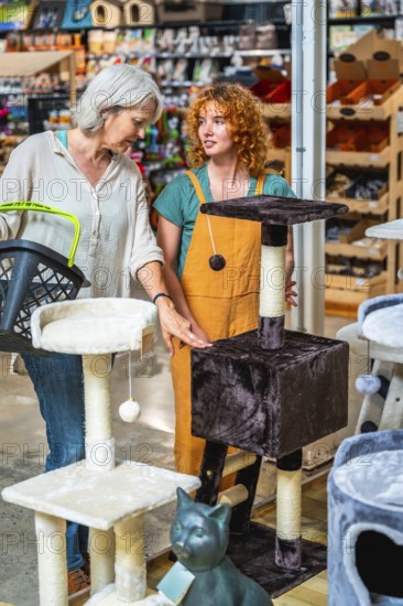 Senior woman holding shopping basket and choosing cat tree with shop assistant help in pet store